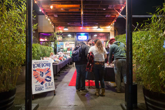 People gathering at the Love Bomb 2017 event entrance, with a colorful poster promoting My Voice Music, and tables displaying items for auction in a vibrant, festive atmosphere.